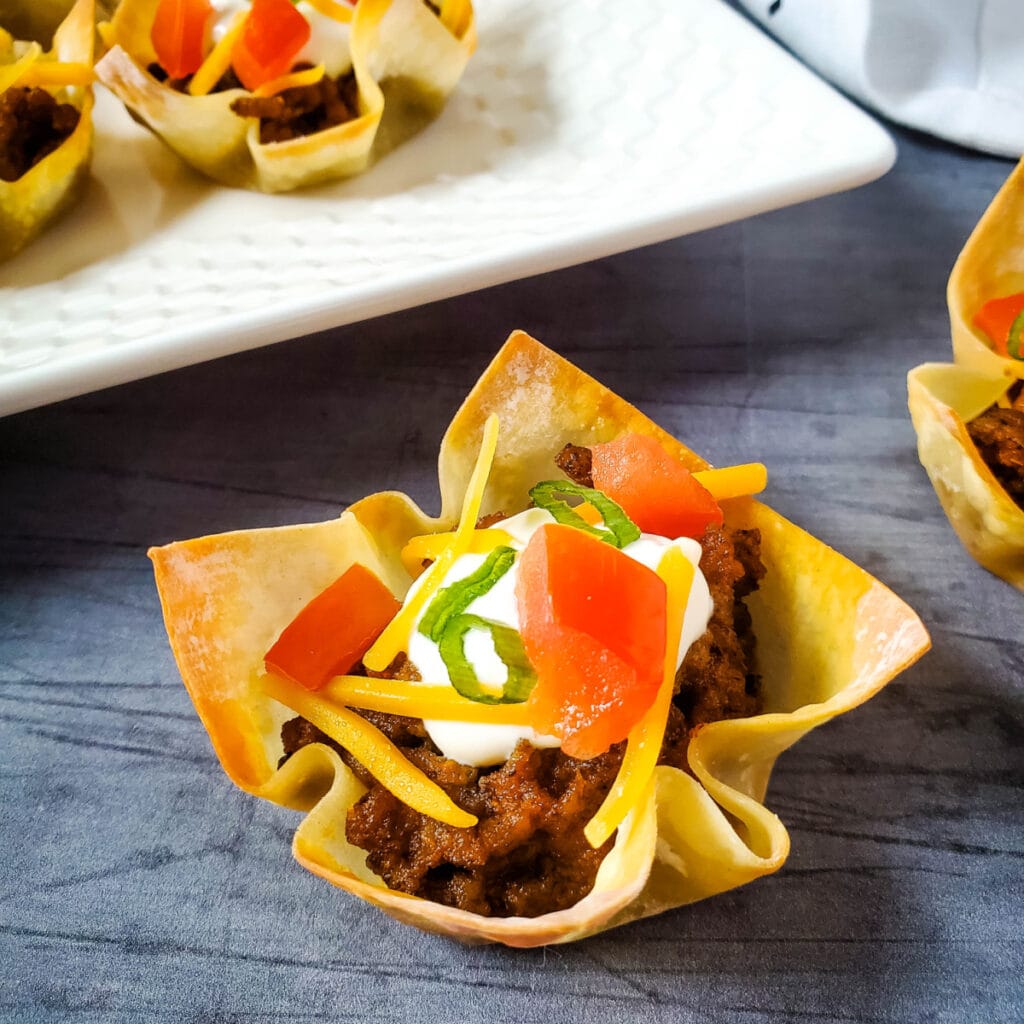 A close-up of a taco cup made with a crispy wonton wrapper, filled with seasoned ground beef, shredded cheese, diced tomatoes, sour cream, and chopped green onions, with more taco cups on a white platter in the background.