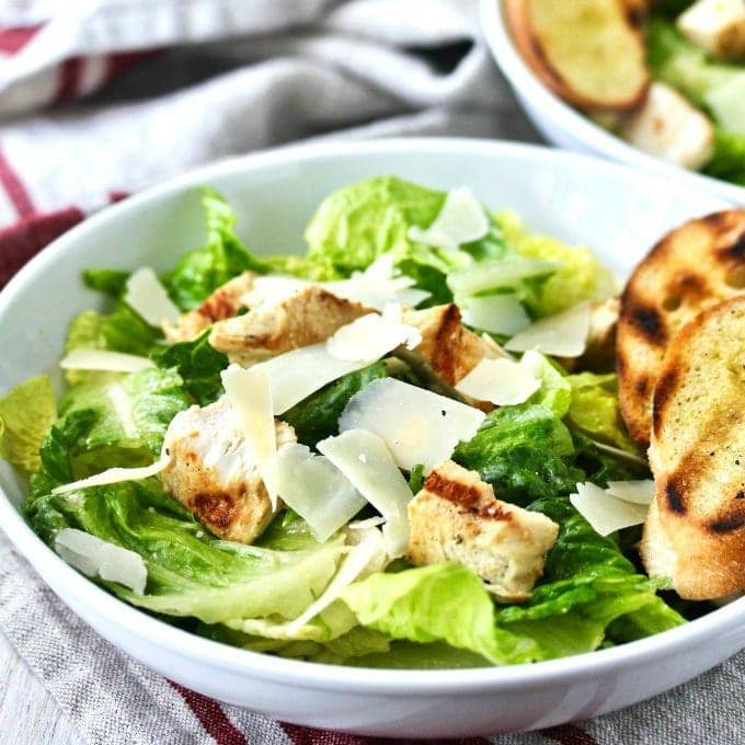 A white bowl of Caesar salad with romaine lettuce, grilled chicken pieces, shaved parmesan cheese, and a slice of toasted bread on the side. A second bowl is partially visible in the background.
