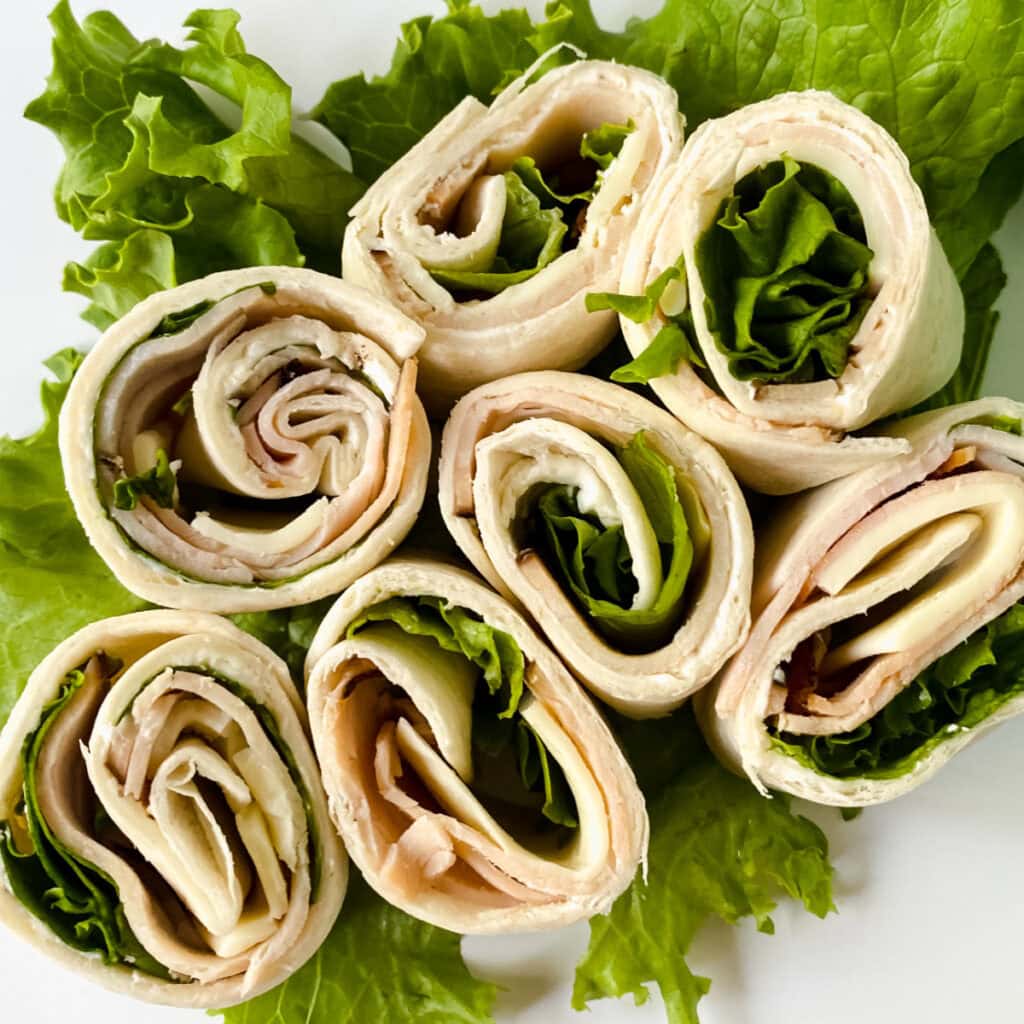Seven turkey and cheese pinwheel wraps with lettuce are arranged on a bed of leafy green lettuce. The wraps are sliced and displayed in a circular pattern, showing their layered fillings.
