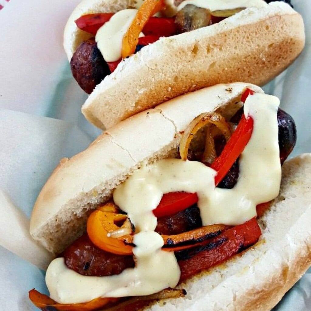 Two grilled sausages in buns topped with sautéed bell peppers and onions, drizzled with creamy sauce, served in a paper-lined basket.