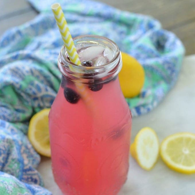 A glass jar filled with pink lemonade, ice, and blueberries, with a yellow-striped straw. Sliced lemons and a patterned cloth are in the background.