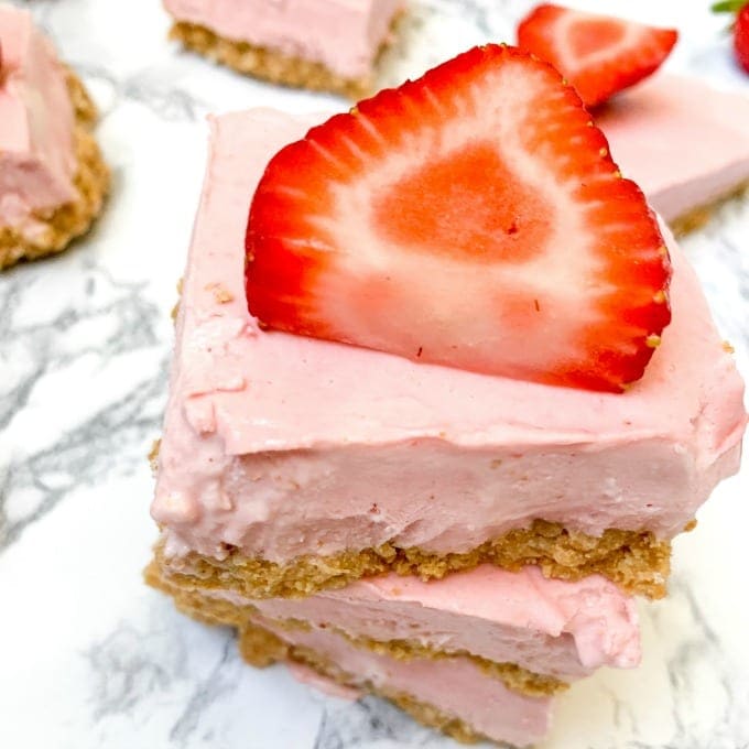 A close-up of two stacked pink strawberry cheesecake bars with a crumbly crust, topped with a fresh strawberry slice on a white marble surface. Other bars are visible in the background.