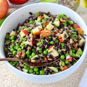 A white bowl filled with wild rice salad containing green peas, chopped apples, pecans, and red onions, with a serving spoon resting inside the bowl.