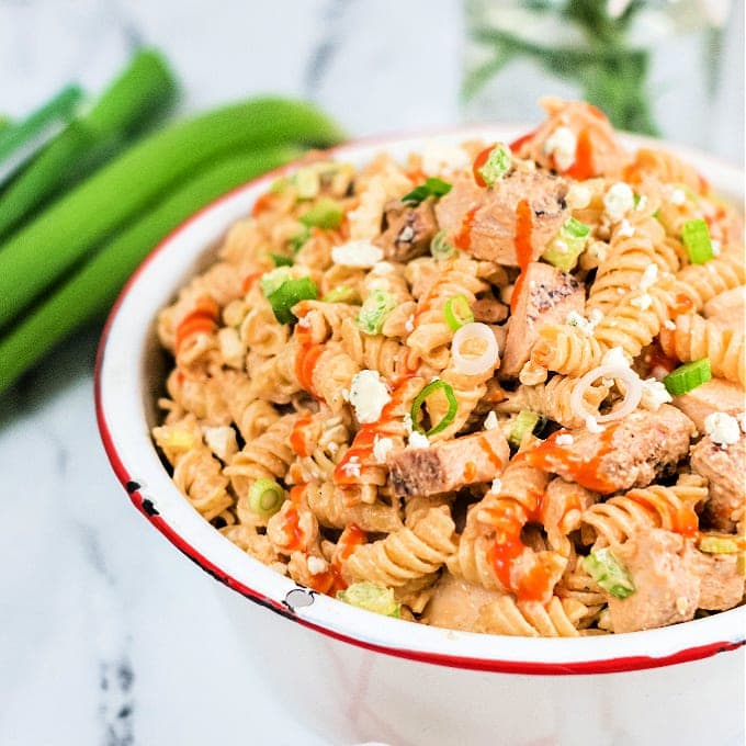 A bowl of rotini pasta salad mixed with grilled chicken pieces, chopped green onions, and drizzled with a red sauce, set on a marble surface with green onions in the background.
