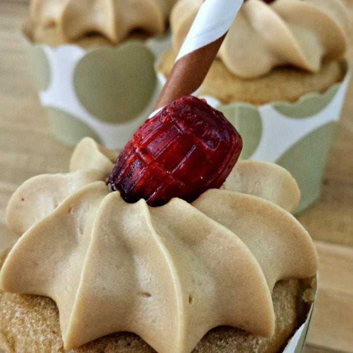 A close-up of a cupcake topped with light brown frosting, a red candy shaped like a barrel, and a paper straw. Two more similarly decorated cupcakes are in the blurred background.