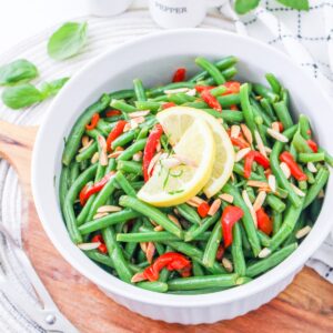 A white bowl filled with green beans, sliced red peppers, and slivered almonds, topped with two lemon slices, sits on a wooden board with fresh herbs and a pepper shaker in the background.
