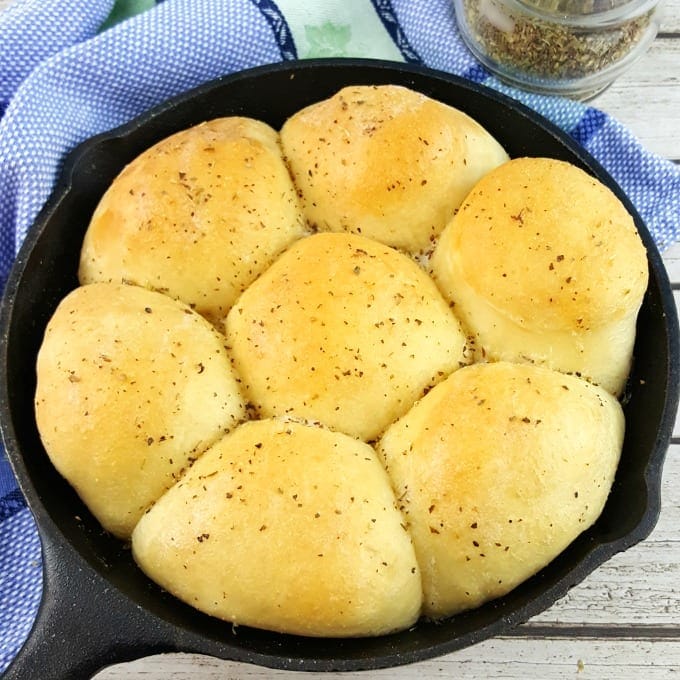 Seven golden-brown dinner rolls seasoned with herbs in a small cast-iron skillet.
