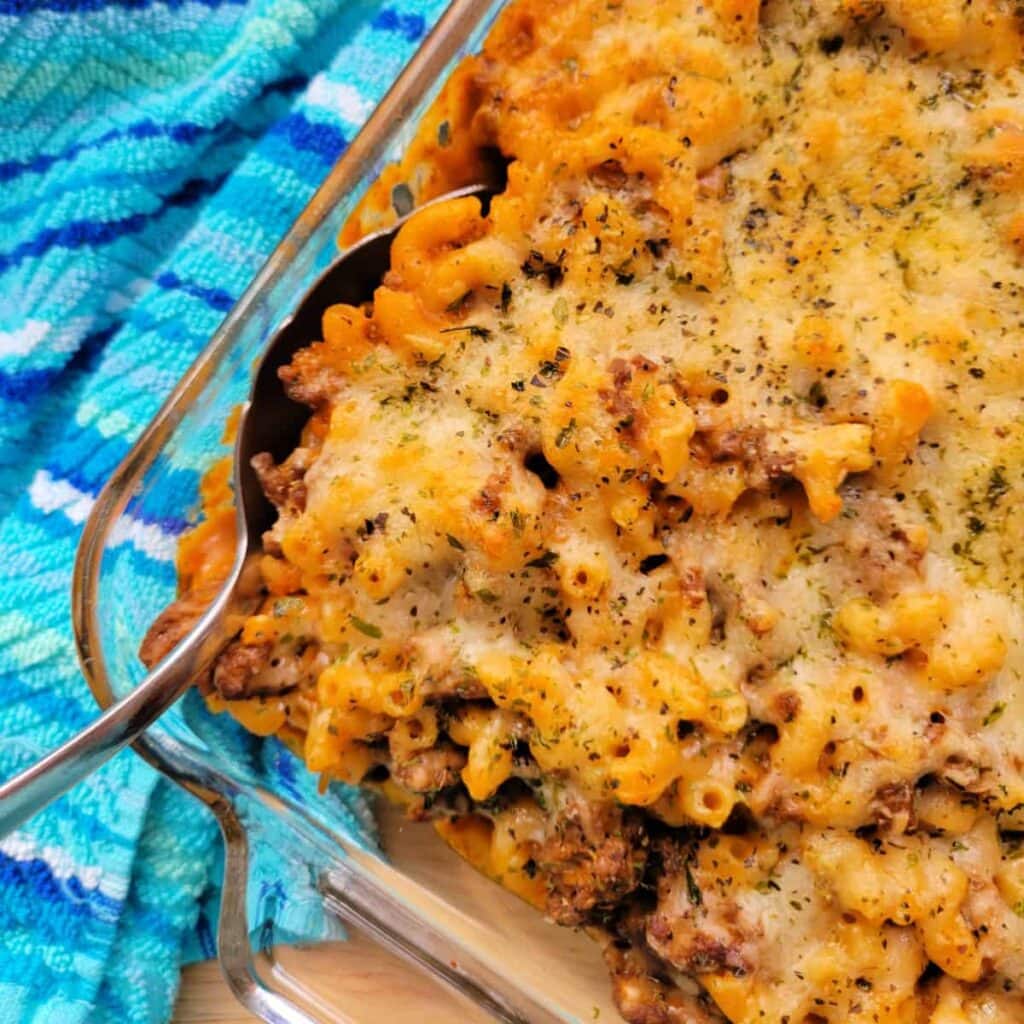 A close-up of a baked pasta casserole with ground meat, melted cheese, and herbs in a glass dish next to a blue patterned towel. A serving spoon is in the dish.