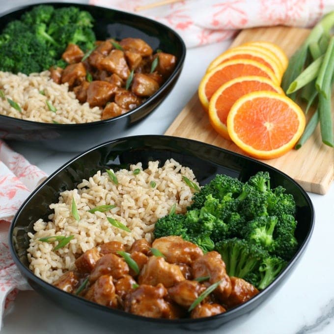30 Minute Chicken Dinners: Two black bowls filled with brown rice, broccoli florets, and orange chicken pieces garnished with green onions. Sliced oranges and green onions are on a wooden board in the background.