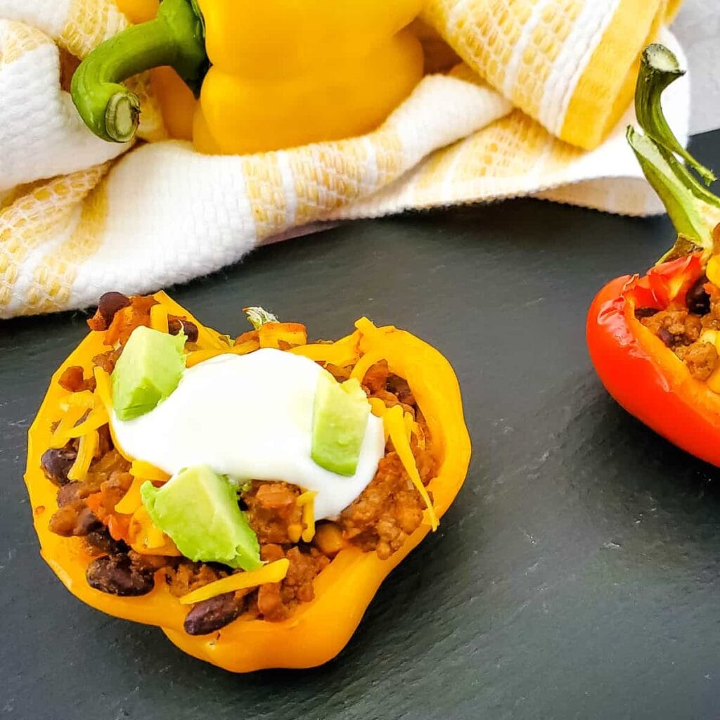 Quick dinner ideas using ground beef: a yellow bell pepper half stuffed with a mixture of ground meat, black beans, and cheese, topped with sour cream and avocado chunks, sits on a dark surface. A red stuffed pepper and a yellow dish towel are in the background.