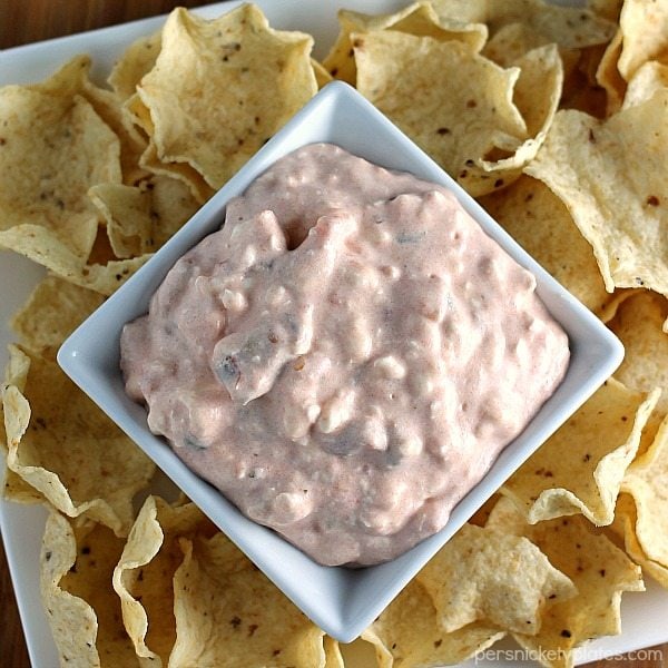 A square white bowl filled with creamy pink dip is surrounded by crispy tortilla chips on a white plate.