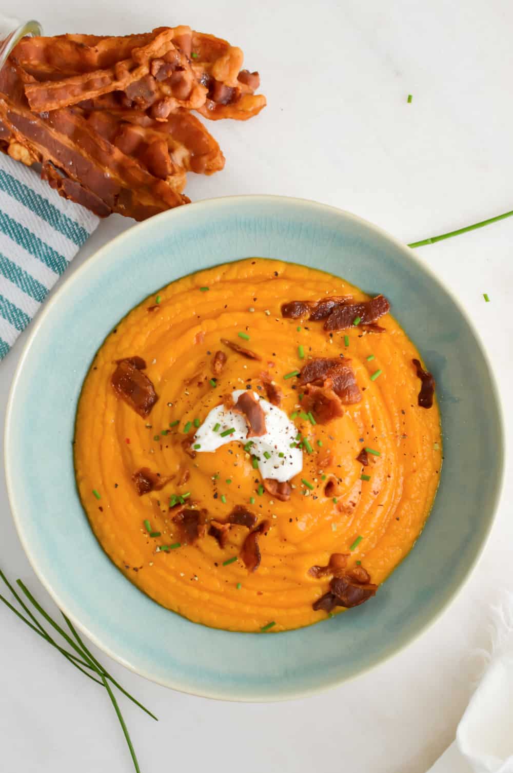 A bowl of creamy orange soup topped with a dollop of sour cream, chopped chives, and crispy bacon bits, with extra bacon strips on the side.