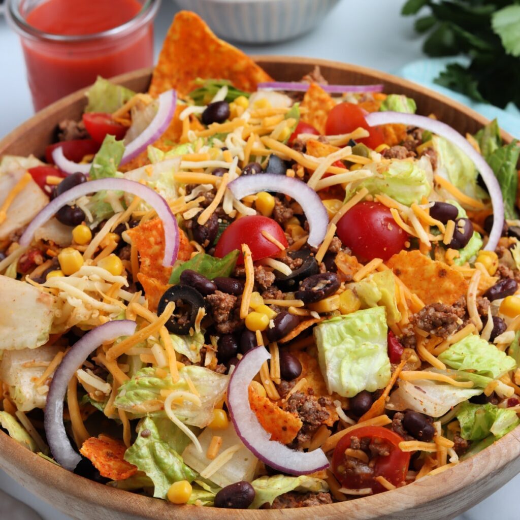 A wooden bowl filled with taco salad, featuring lettuce, cherry tomatoes, black beans, corn, shredded cheese, red onion slices, ground beef, and tortilla chips. A glass of red dressing is visible in the background.