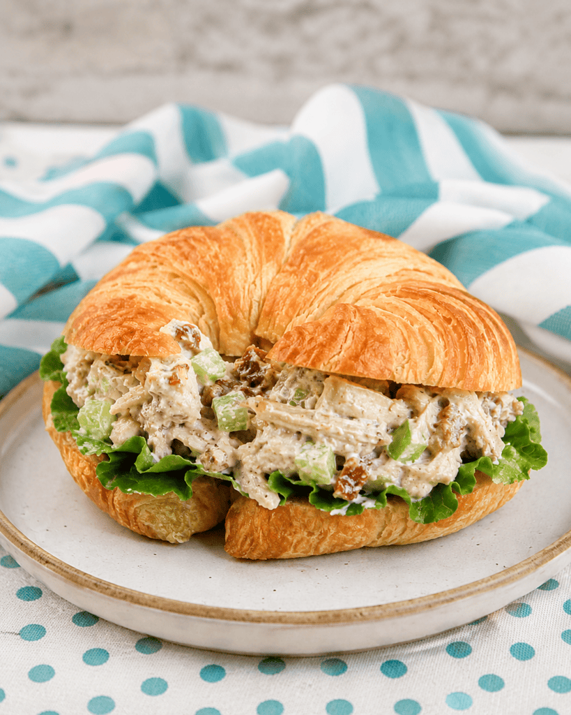 A croissant sandwich filled with Rotisserie Chicken Salad, lettuce, and chopped vegetables sits on a plate. A blue and white striped cloth is in the background.
