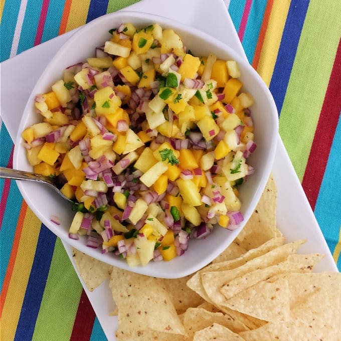 A white bowl of fresh salsa with diced mango, pineapple, red onion, and cilantro sits next to tortilla chips on a white plate. The background is a colorful striped tablecloth.