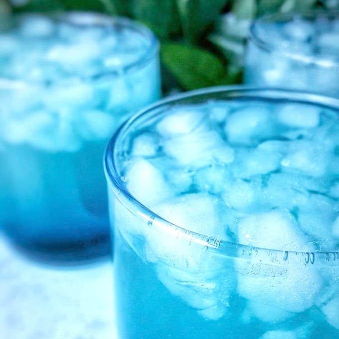 Three clear glasses filled with blue liquid and ice cubes, with condensation on the outside. The background is blurred with hints of green leaves.