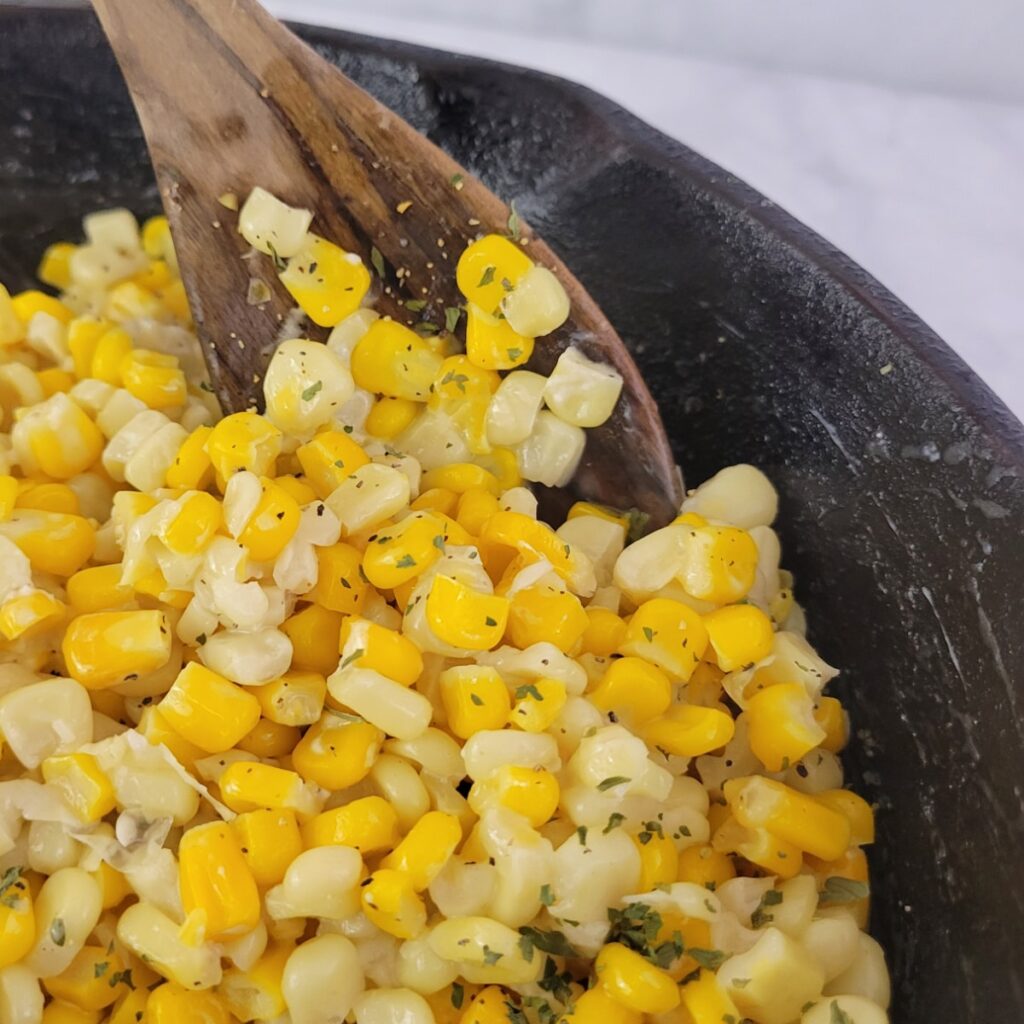 A wooden spoon stirring a mixture of yellow and white corn kernels in a black cast-iron skillet.