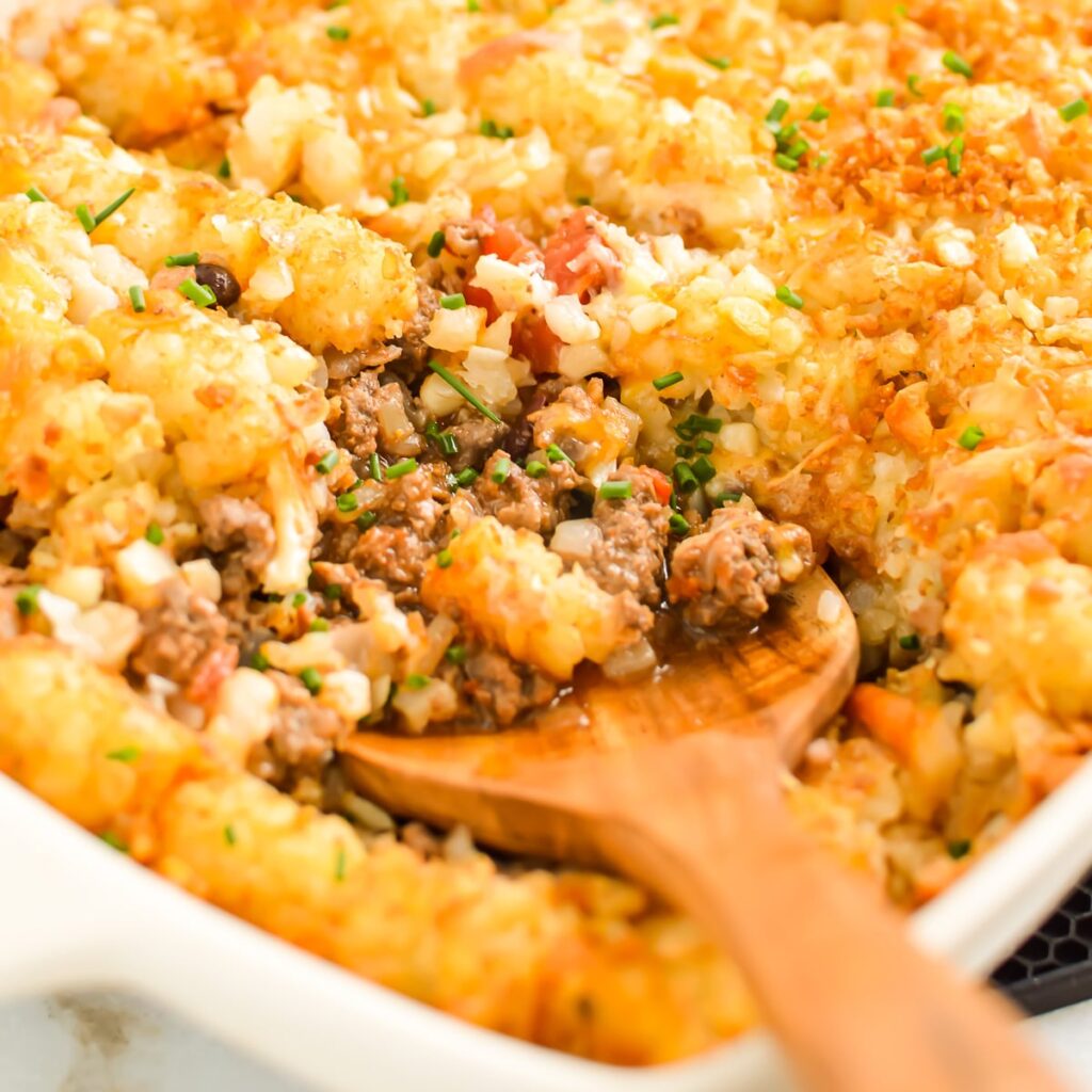 Ground beef dinner recipes: a close-up of a casserole dish filled with tater tot hotdish; a wooden spoon scoops out a portion, revealing ground beef, melted cheese, and crispy tater tots, garnished with chopped herbs.
