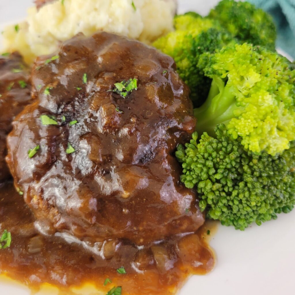 Easy ground beef dinner recipes: close-up of Salisbury steak covered in brown onion gravy, served with a side of steamed broccoli and mashed potatoes on a white plate.
