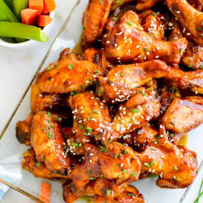 A glass dish filled with glazed chicken wings garnished with sesame seeds and chopped herbs, with a side of celery and carrot sticks in the corner.