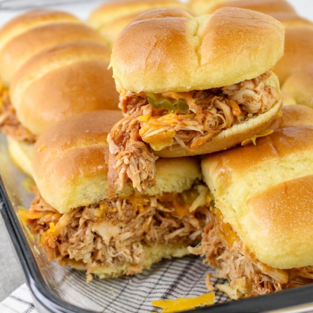 A close-up of several pulled chicken sliders with shredded chicken and melted cheese on soft buns, stacked in a glass baking dish. One slider is stacked on top, slightly tilted to show the filling.
