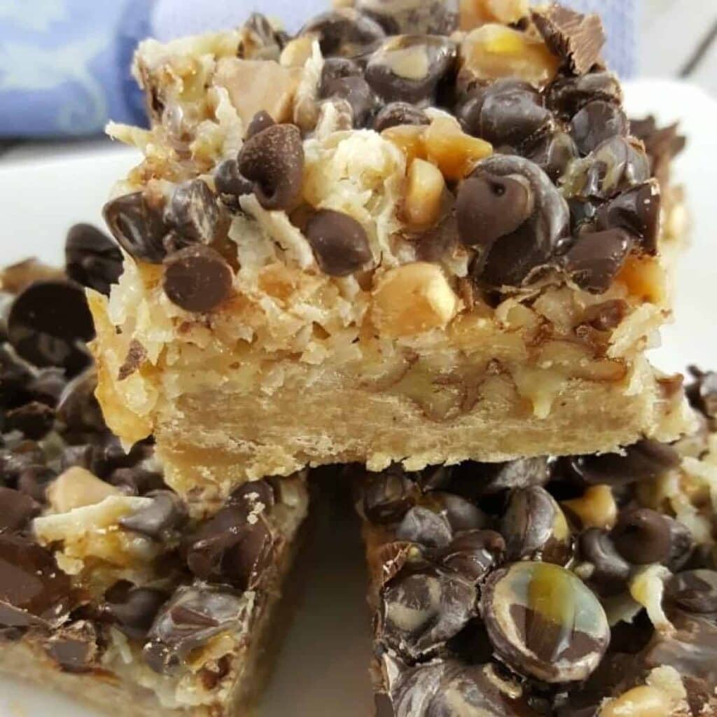 Close-up of dessert bars stacked on a plate, showing layers of cookie crust, coconut, chopped nuts, and generous toppings of chocolate chips and butterscotch chips.