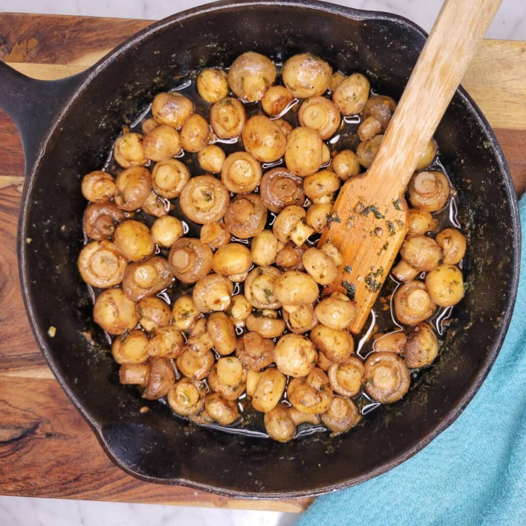 Cast iron skillet filled with sautéed brown mushrooms in a garlic butter sauce, with a wooden spatula resting inside.