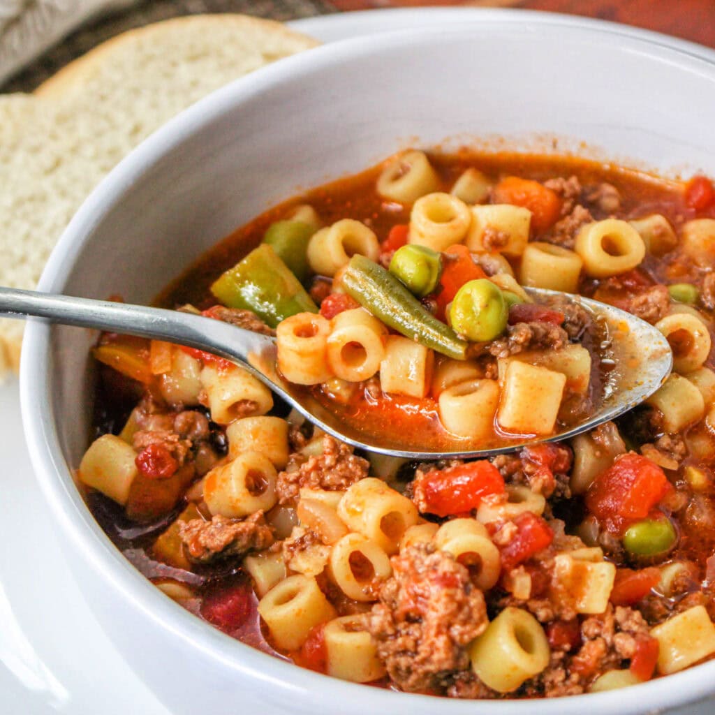 Ground beef dinner ideas: a close-up of a bowl of minestrone soup with small tube pasta, ground meat, peas, green beans, carrots, and tomatoes. A spoon is scooping up the hearty mixture. Sliced bread is visible in the background.