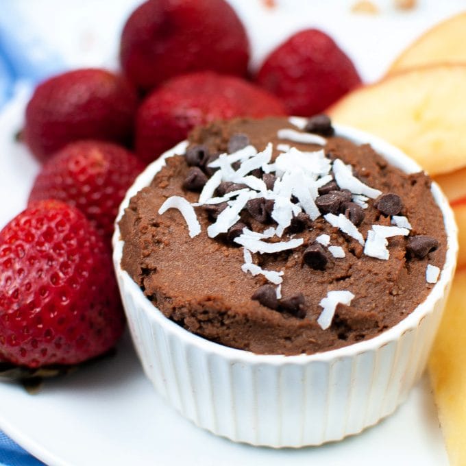 A small white ramekin filled with chocolate dip, topped with shredded coconut and chocolate chips, is surrounded by fresh strawberries and sliced apples on a white plate.