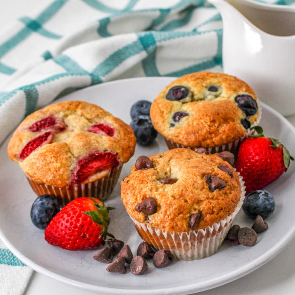 A plate with three muffins—one with strawberries, one with blueberries, and one with chocolate chips—surrounded by fresh strawberries, blueberries, and chocolate chips, with a white-and-turquoise towel in the background.