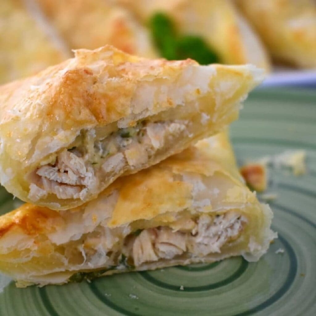 Close-up of two flaky, golden-brown puff pastry triangles filled with shredded chicken, stacked on a green plate. The pastry looks crisp and the filling is visible inside.
