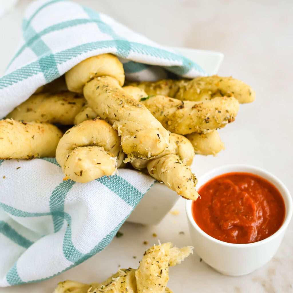 Italian Breadsticks with herbs and cheese in a basket with a kitchen cloth next to marinara sauce.