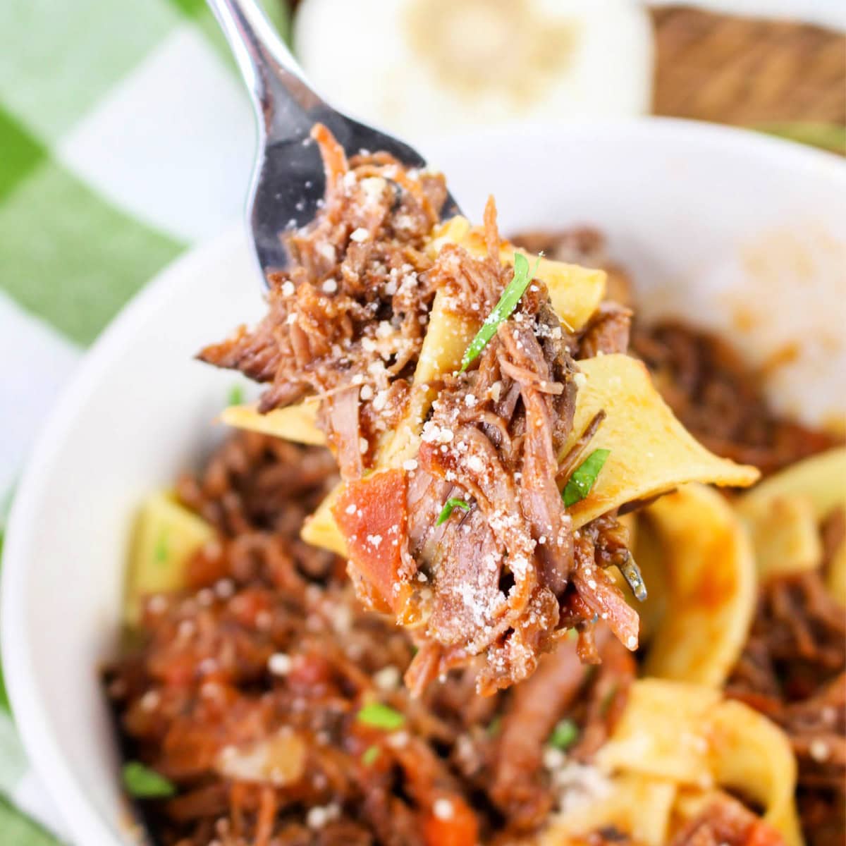 A close-up of a fork holding pasta with beef ragu tomato sauce, and grated cheese above a bowl filled with more pasta and sauce.
