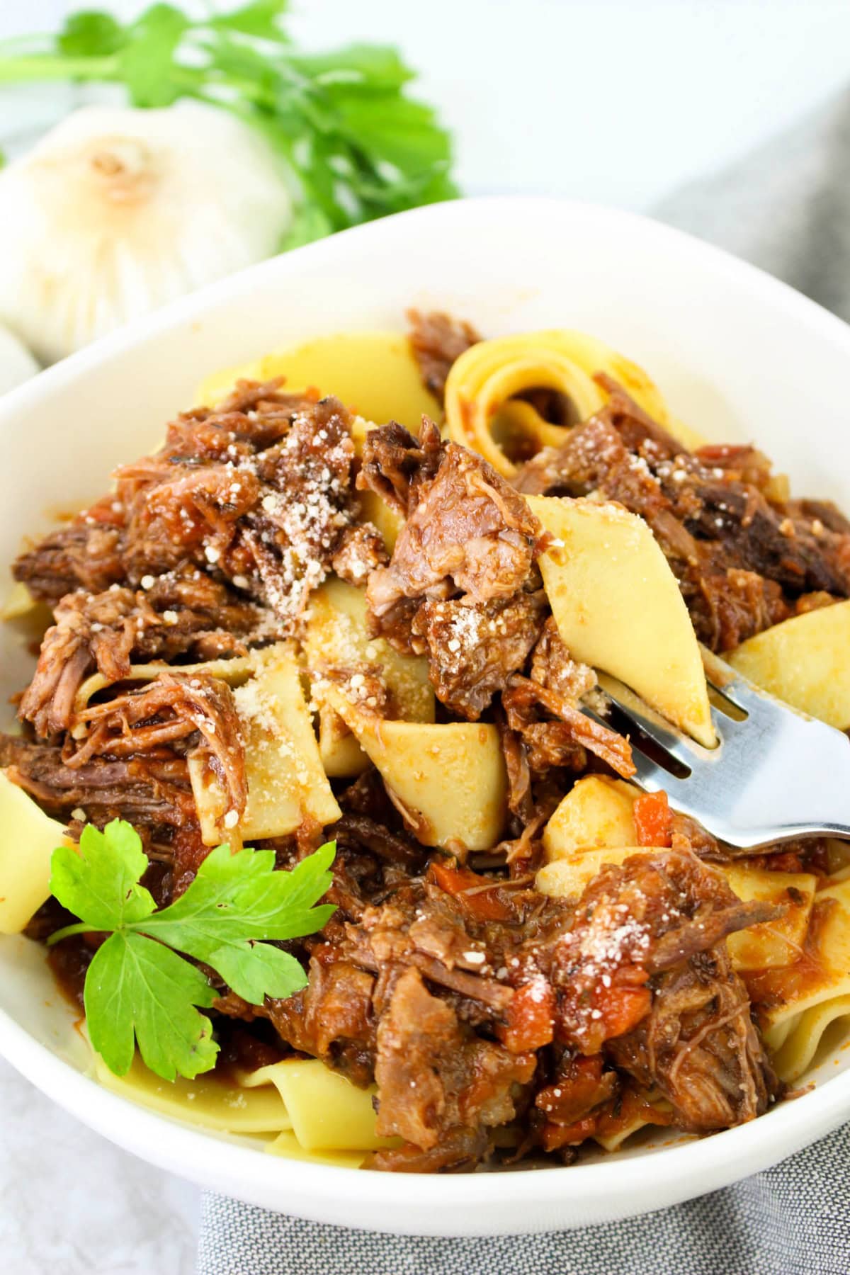 A bowl of pappardelle pasta topped with slow-cooked shredded beef ragu, garnished with grated cheese and fresh parsley. A fork rests in the bowl, and garlic and parsley are in the background.