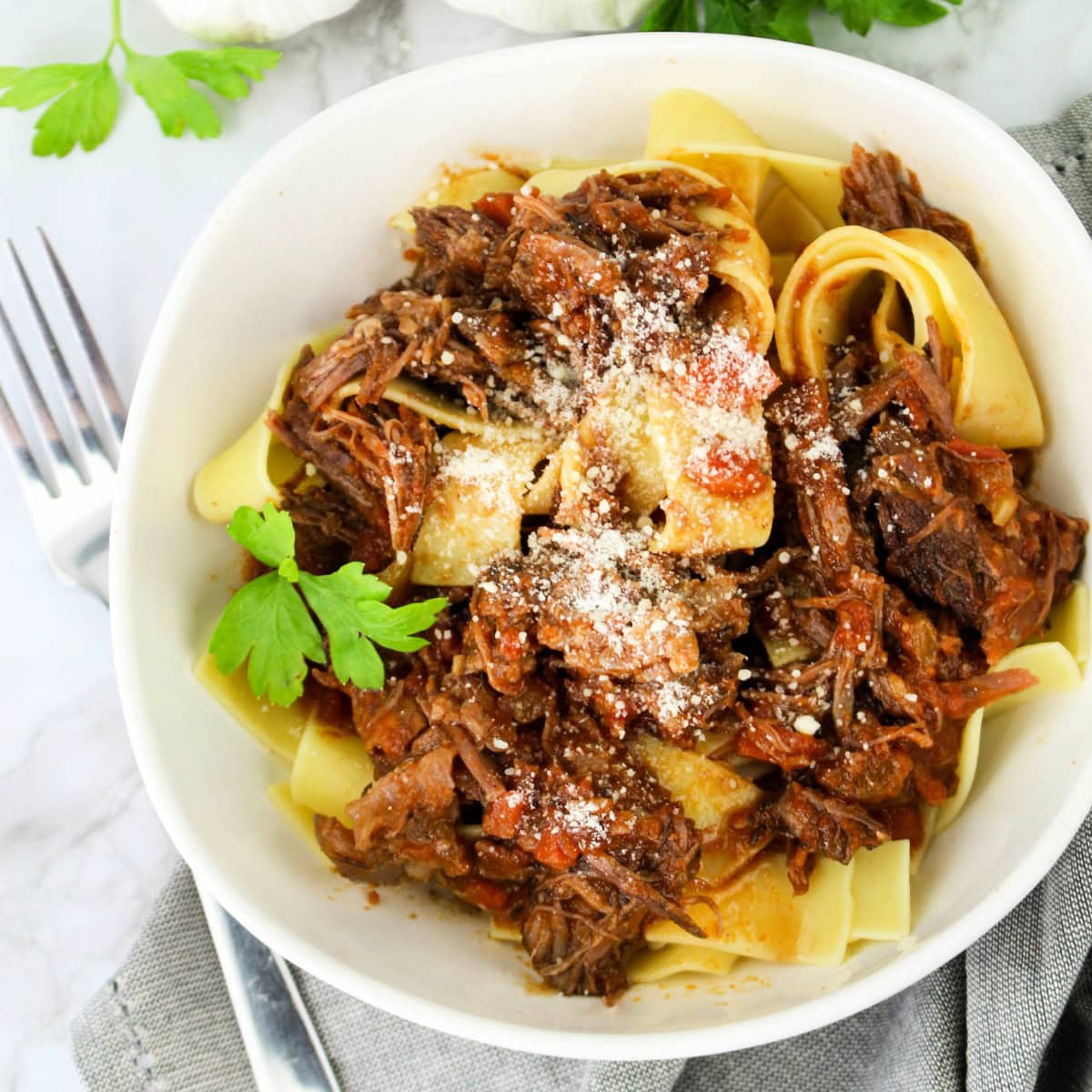 A white bowl filled with wide pasta noodles topped with shredded beef ragu, garnished with grated cheese and fresh parsley. A fork and napkin sit beside the bowl on a marble surface.