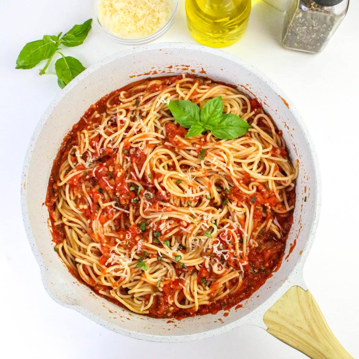 A white pan filled with spaghetti mixed in tomato sauce, garnished with fresh basil and grated cheese. Nearby are a basil sprig, grated cheese, olive oil, and a pepper grinder on a white surface.