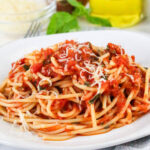 A plate of spaghetti topped with tomato sauce and grated cheese, garnished with herbs, sits on a white dish. In the background, there are hints of a cheese bowl, green basil, and a yellow olive oil bottle.