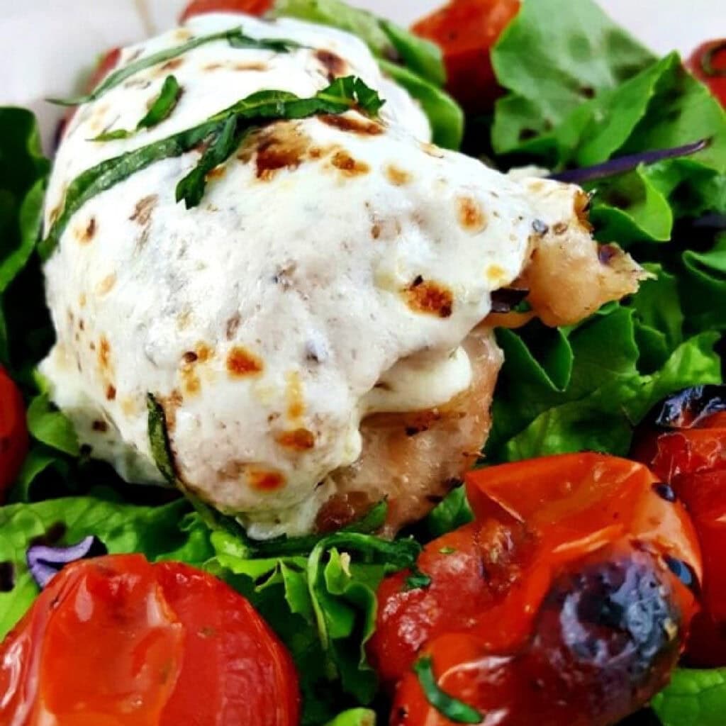 A close-up of a baked chicken breast topped with melted cheese and herbs, served on a bed of fresh greens with roasted cherry tomatoes.