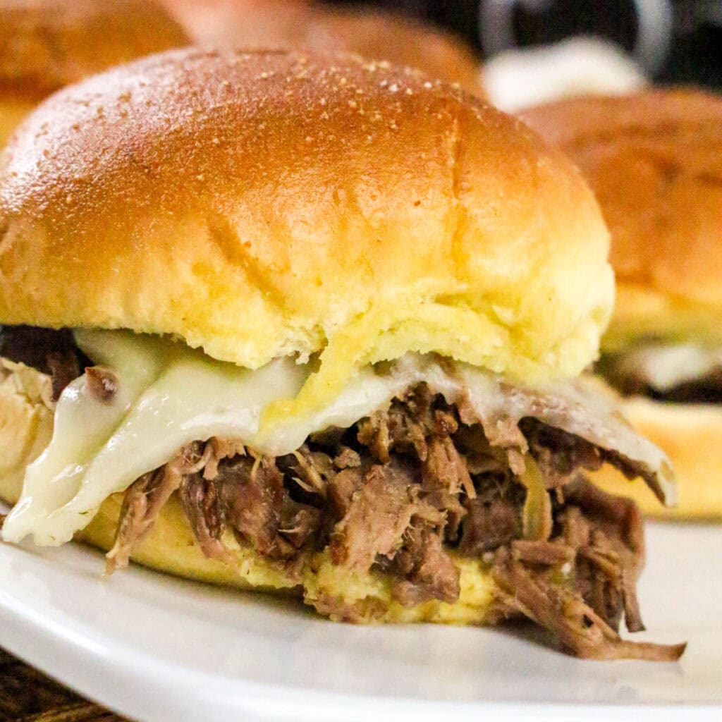A close-up of a sandwich with shredded beef and melted cheese on a soft, golden bun, served on a white plate. Another similar sandwich is visible in the background.