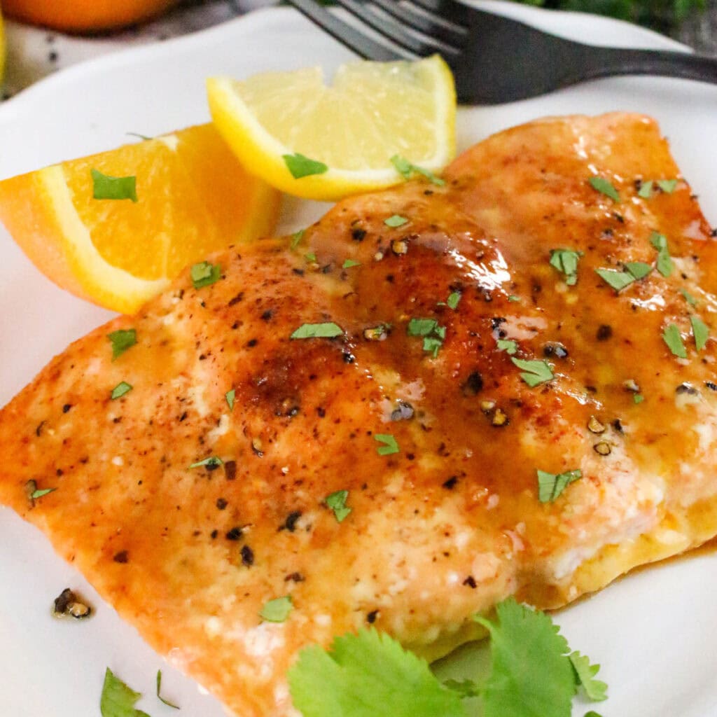 A fillet of baked citrus glazed salmon topped with herbs and cracked pepper, garnished with fresh orange and lemon wedges, on a white plate.