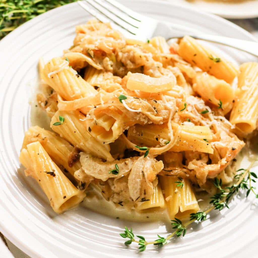 A plate of rigatoni pasta topped with creamy caramelized onions and fresh thyme, served on a white dish with a fork in the background.