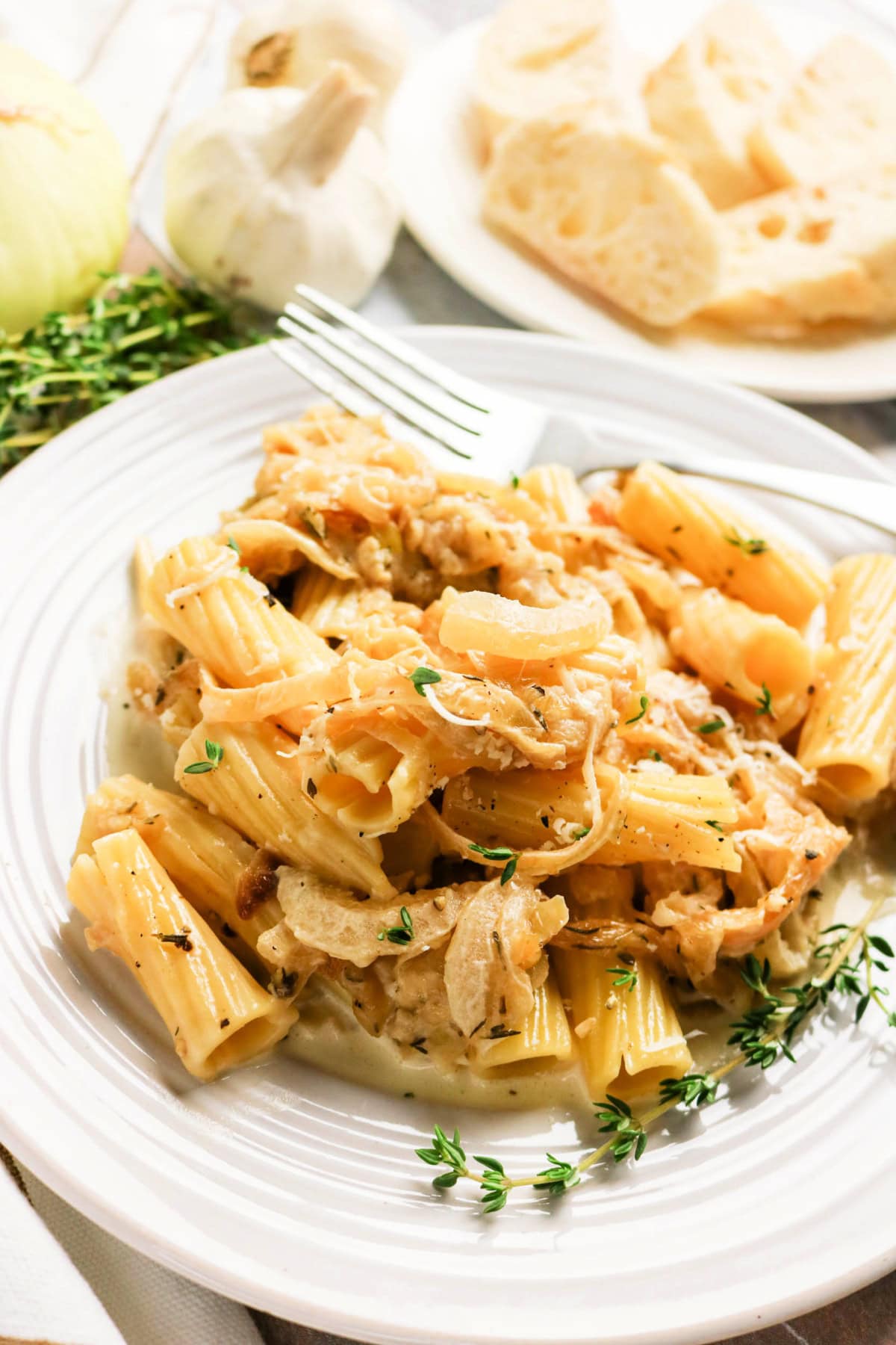 A plate of rigatoni pasta topped with creamy sauce, caramelized onions, and fresh thyme. In the background, there’s a plate of sliced bread, a garlic bulb, and fresh herbs.
