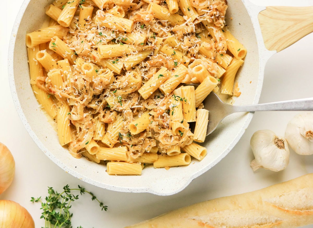 A skillet filled with creamy rigatoni pasta topped with grated cheese and herbs. Surrounding the skillet are fresh garlic, onions, thyme, and part of a baguette on a white surface.