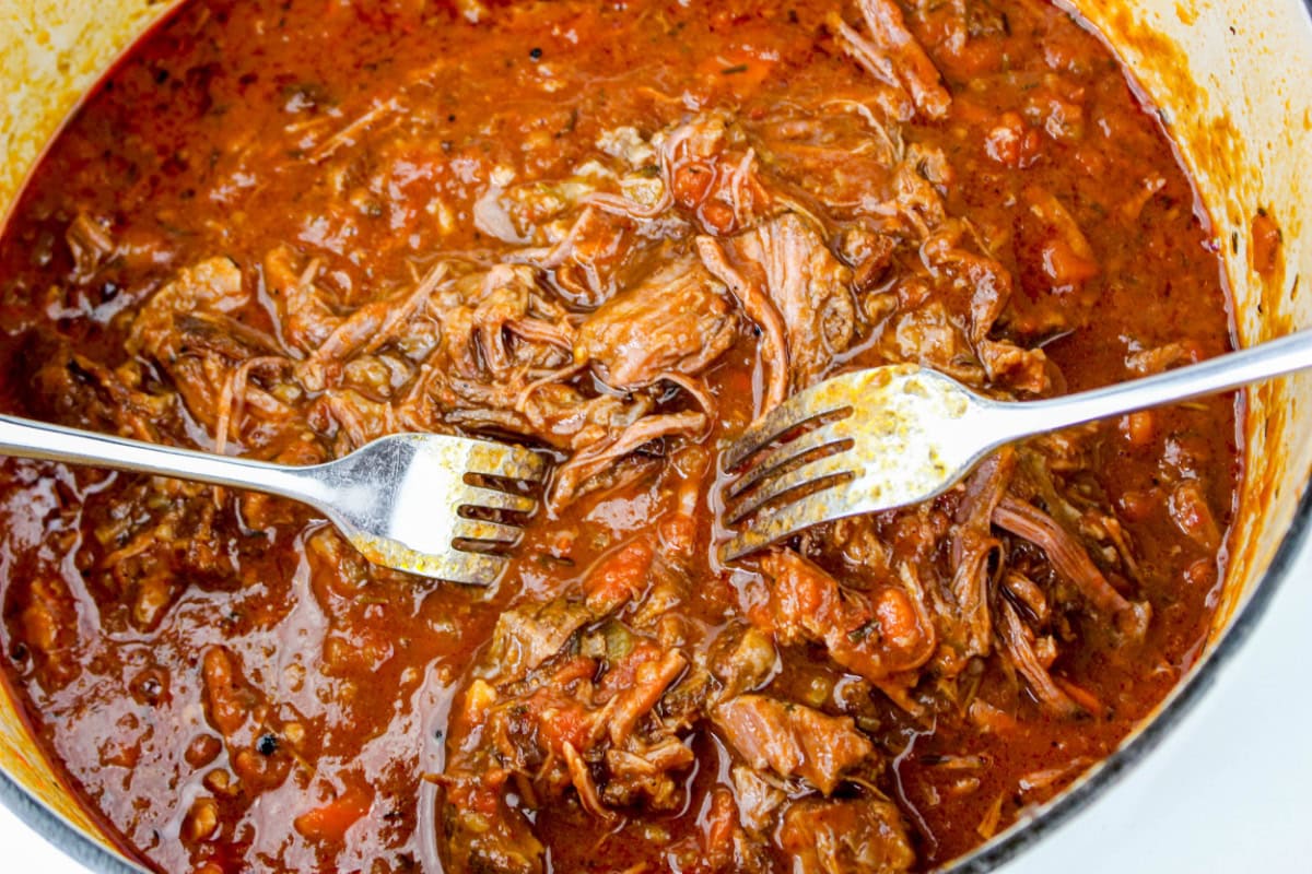 Two metal forks are shredding tender, slow-cooked beef in a pot of rich, red sauce.