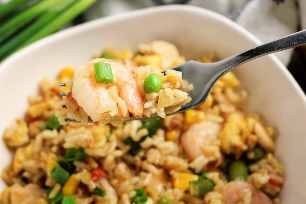 A close-up of a fork holding a bite of chicken and shrimp fried rice with peas, green onions, egg, and vegetables, above a bowl filled with the same dish.