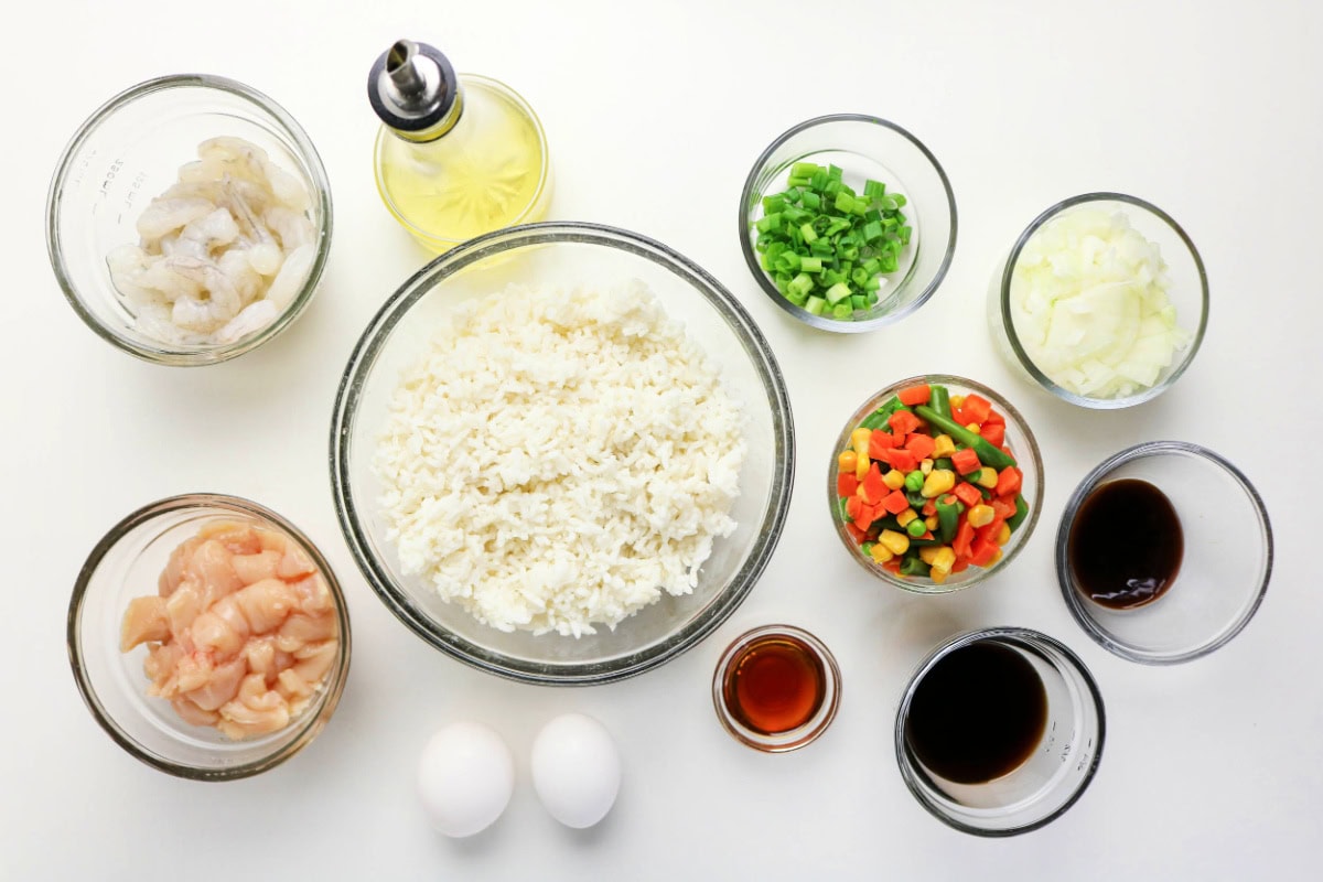 Top-down view of bowls containing ingredients for Shrimp and Chicken Fried Rice: cooked rice, raw shrimp, diced chicken, chopped green onions, diced white onion, mixed vegetables, soy sauce, another dark sauce, oil, and two eggs on a white surface.