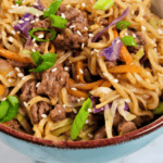 A close-up of a bowl filled with stir-fried noodles, ground beef, green onions, shredded carrots, cabbage, and sesame seeds. The Beef and Ramen Noodles dish is served in a turquoise bowl.