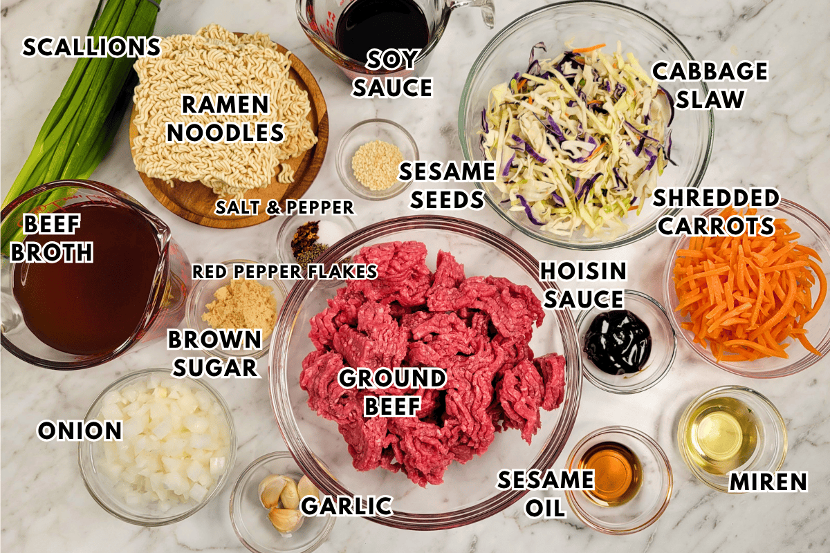 A top-down view of ingredients for a spicy beef ramen dish, including raw ground beef, ramen noodles, beef broth, chopped scallions, diced onion, garlic, sesame seeds, soy sauce, hoisin sauce, sesame oil, mirin, brown sugar, red pepper flakes, cabbage slaw, and shredded carrots.