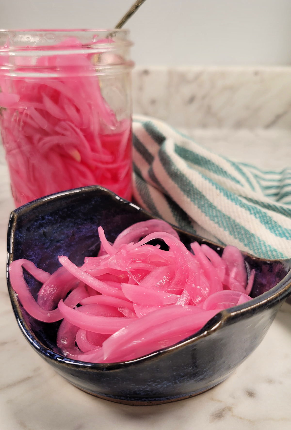 A close-up of pickled red onions in a dark ceramic bowl, with a jar of more pickled onions and a striped kitchen towel in the background on a marble countertop.