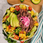 A colorful salad bowl with shredded beef, sliced avocado, pickled red onions, grilled corn, jalapeño, cherry tomatoes, bell peppers, cucumber, and lime wedges on a marble surface with a striped cloth nearby.